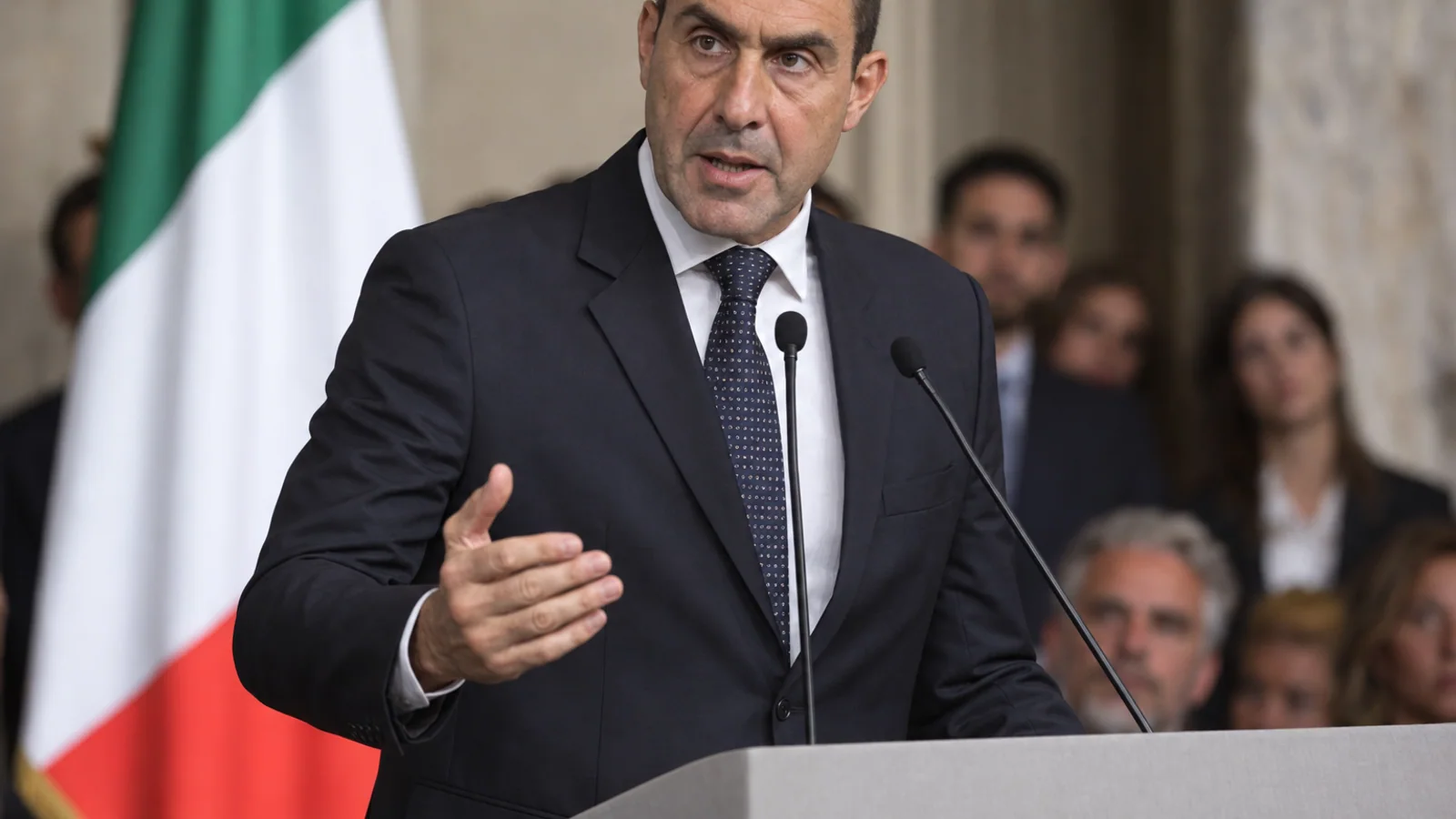Roberto Vannacci speaks at a podium during a political event, with blurred Italian tricolour colours in the background.