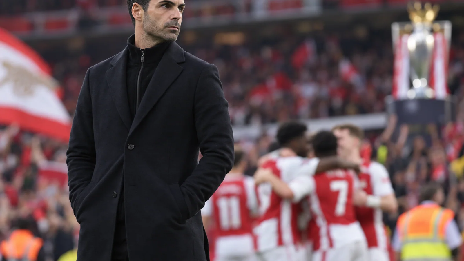 Mikel Arteta stands on the touchline during a football match, with a stadium crowd and a blurred trophy in the background.